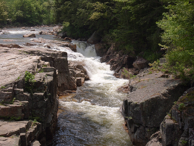 [The stream comes toward the camera down the left side and then in the middle back of the image it drops several feet toward the right in multiple places creating lots of white water. It then proceeds straight toward the camera with large hunks of rock on either side of the water flow.]